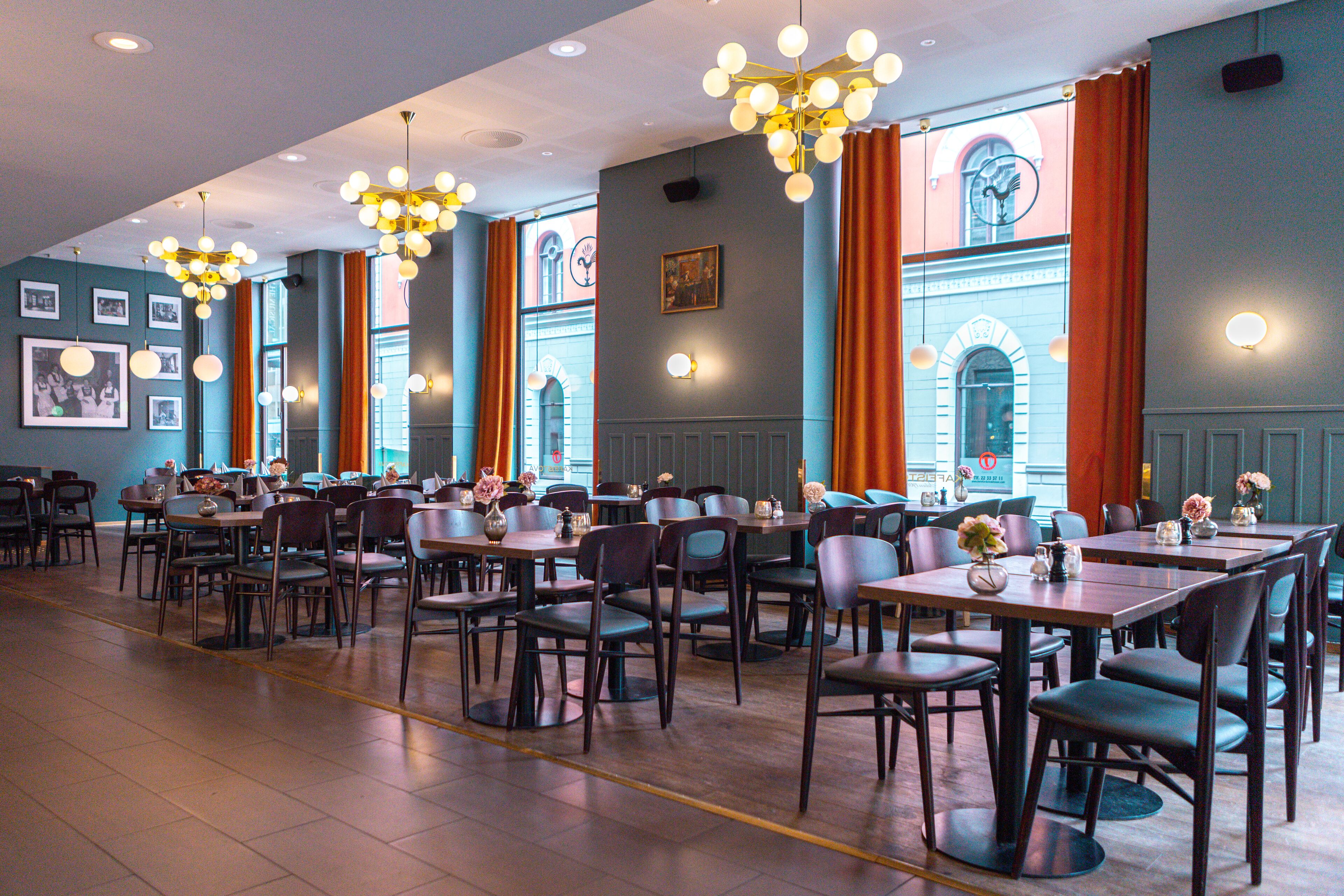 Un restaurant doté de nombreuses tables et chaises, de grandes fenêtres avec des rideaux et de luminaires suspendus.