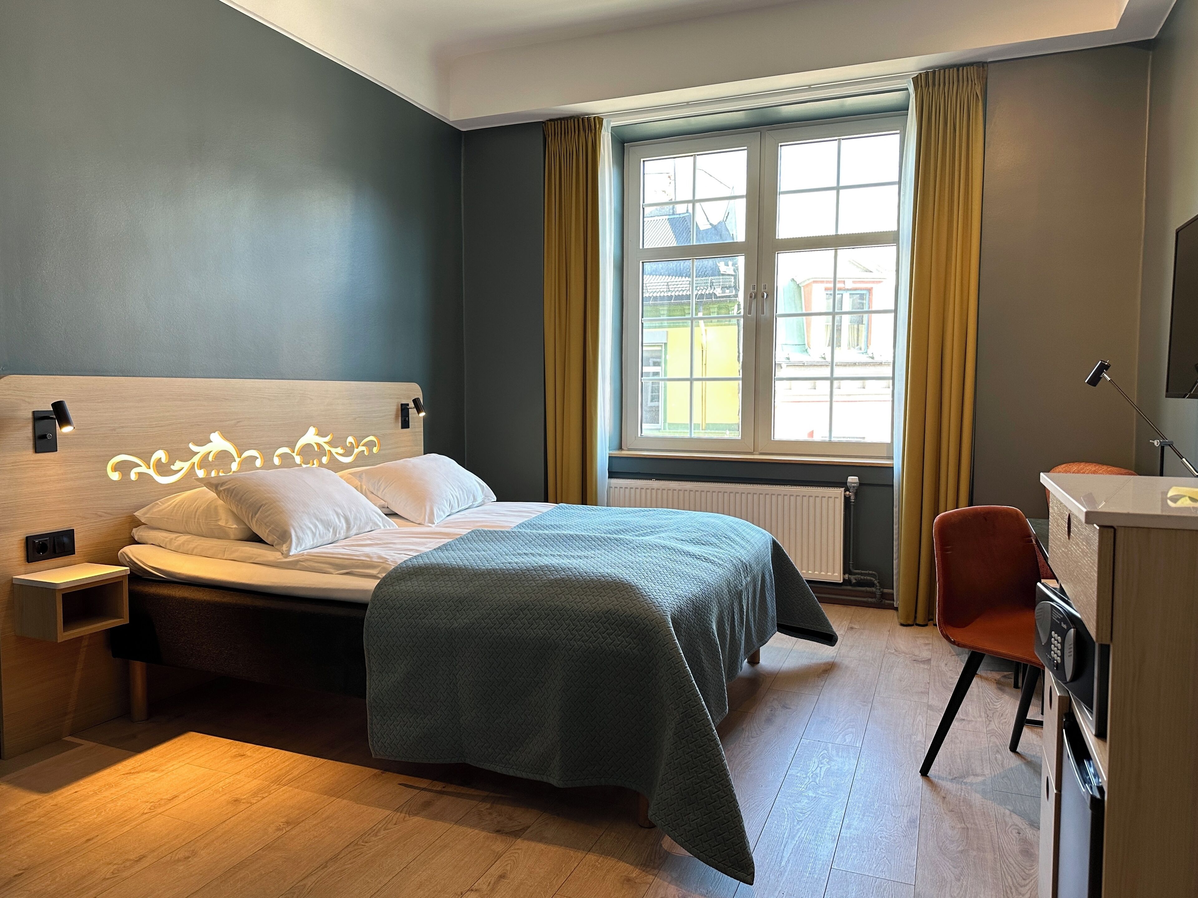A modern hotel room with a bed, bedside table, and a desk with a chair.