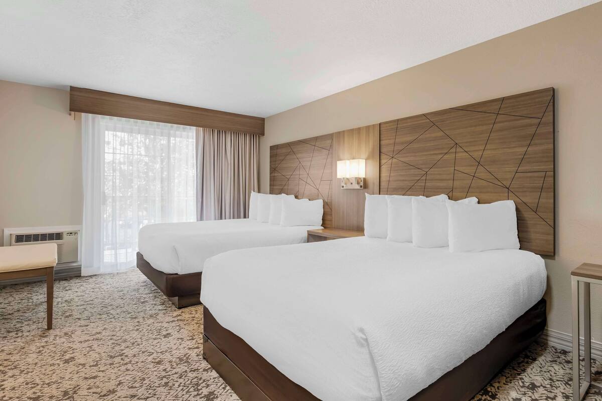 A hotel room with two beds, a large window with curtains, a bedside table, and a wall-mounted lamp.