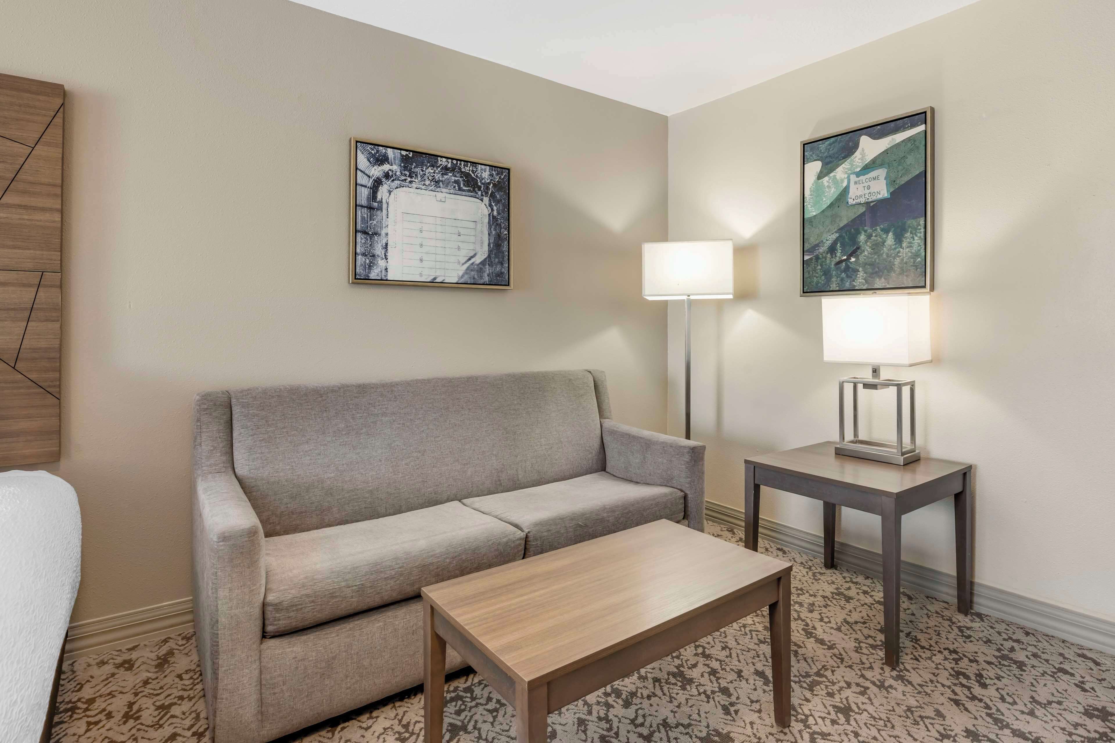 A hotel room with a grey sofa, a wooden coffee table, a lamp on a side table, and two framed pictures on the walls.