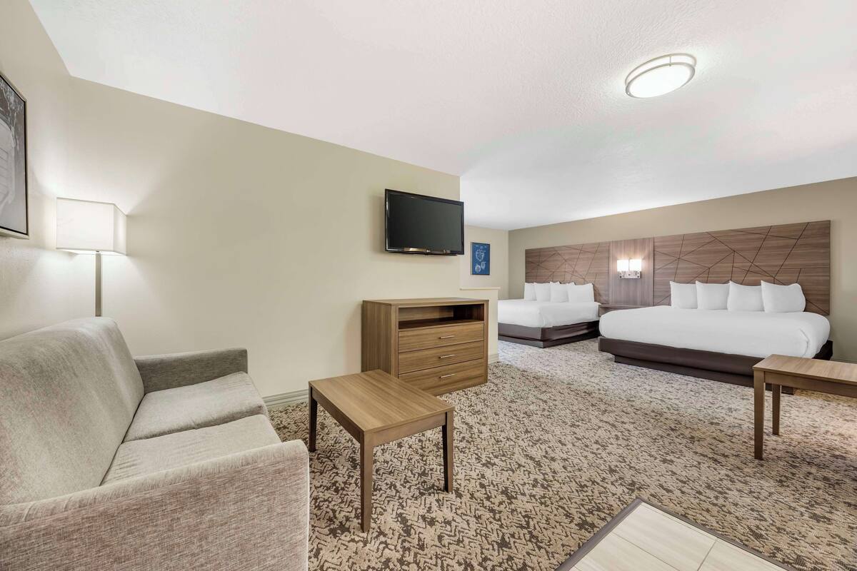 A hotel room with a sofa, a small wooden table, a TV, a bed with two pillows, and a headboard with a geometric pattern.