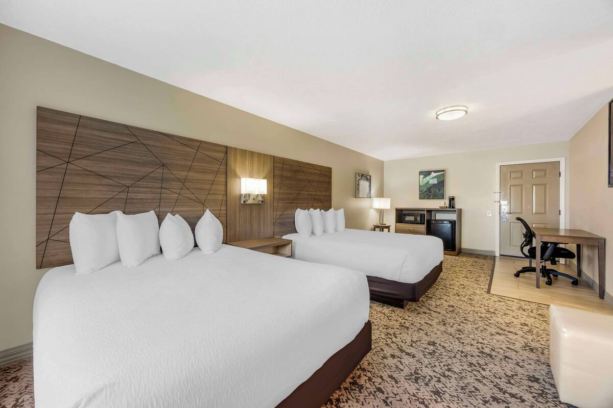A hotel room with two beds, a desk, a chair, a lamp, and a small kitchenette.