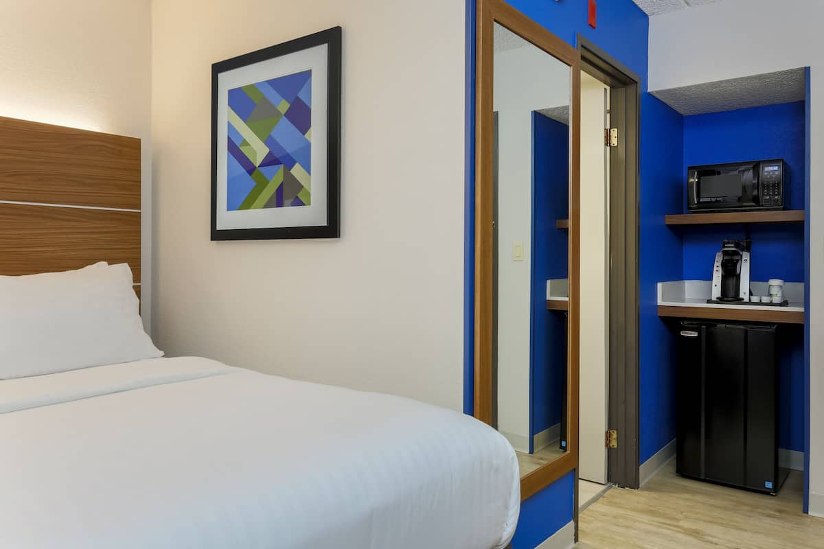 A hotel room with a bed, a framed abstract painting, a microwave, a coffee maker, and a refrigerator.