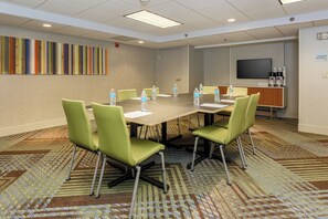 Une salle de conférence avec une grande table, des chaises vertes, des bouteilles d’eau et un téléviseur fixé au mur.