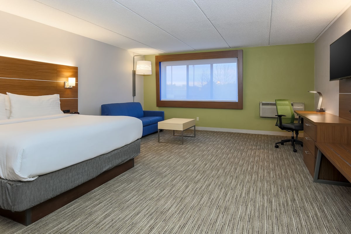 A hotel room with a bed, a desk, a chair, a small table, a lamp, and a window.