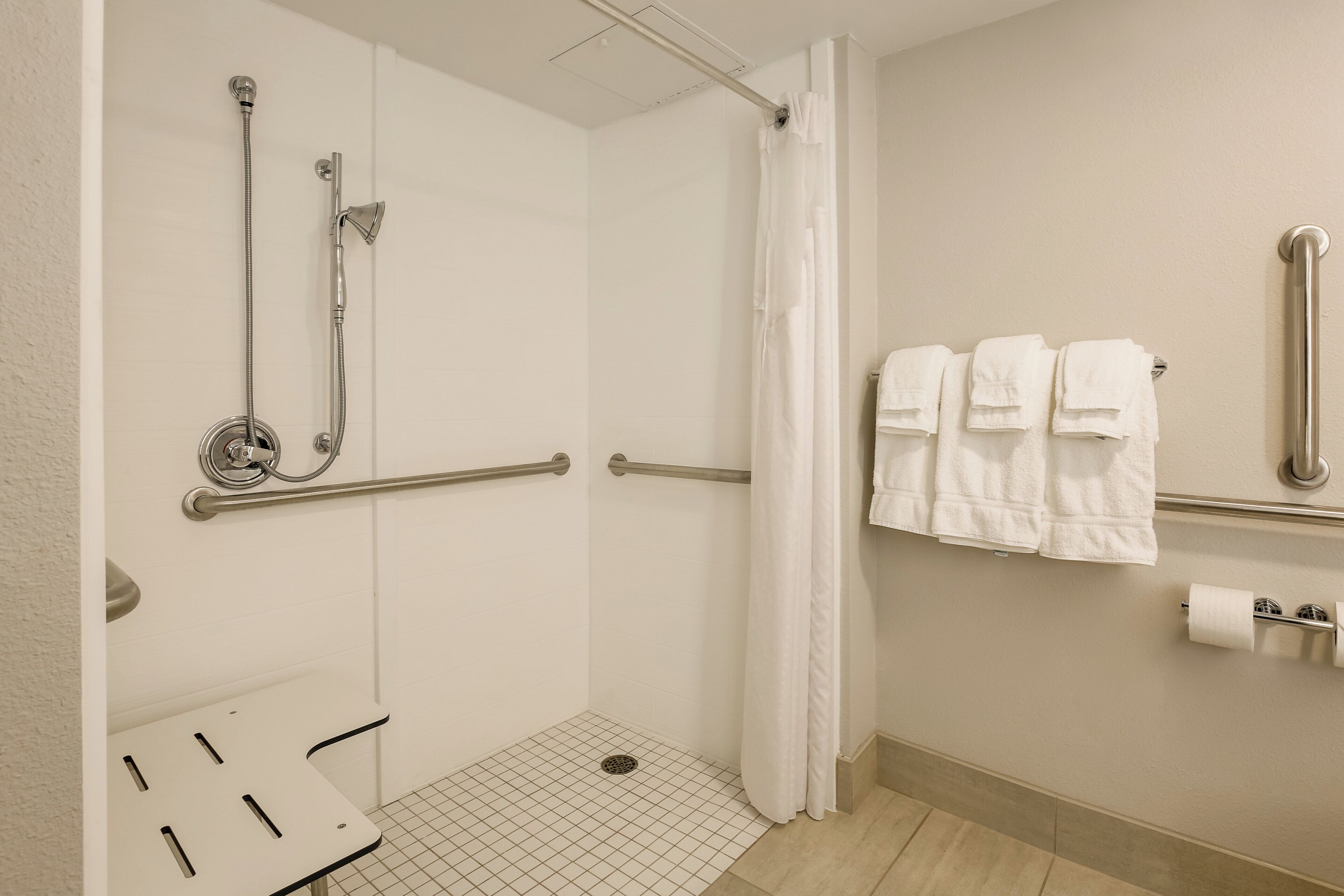 Ducha accesible para silla de ruedas con barras de apoyo, suelo de baldosas blancas y un perchero con toallas dobladas.