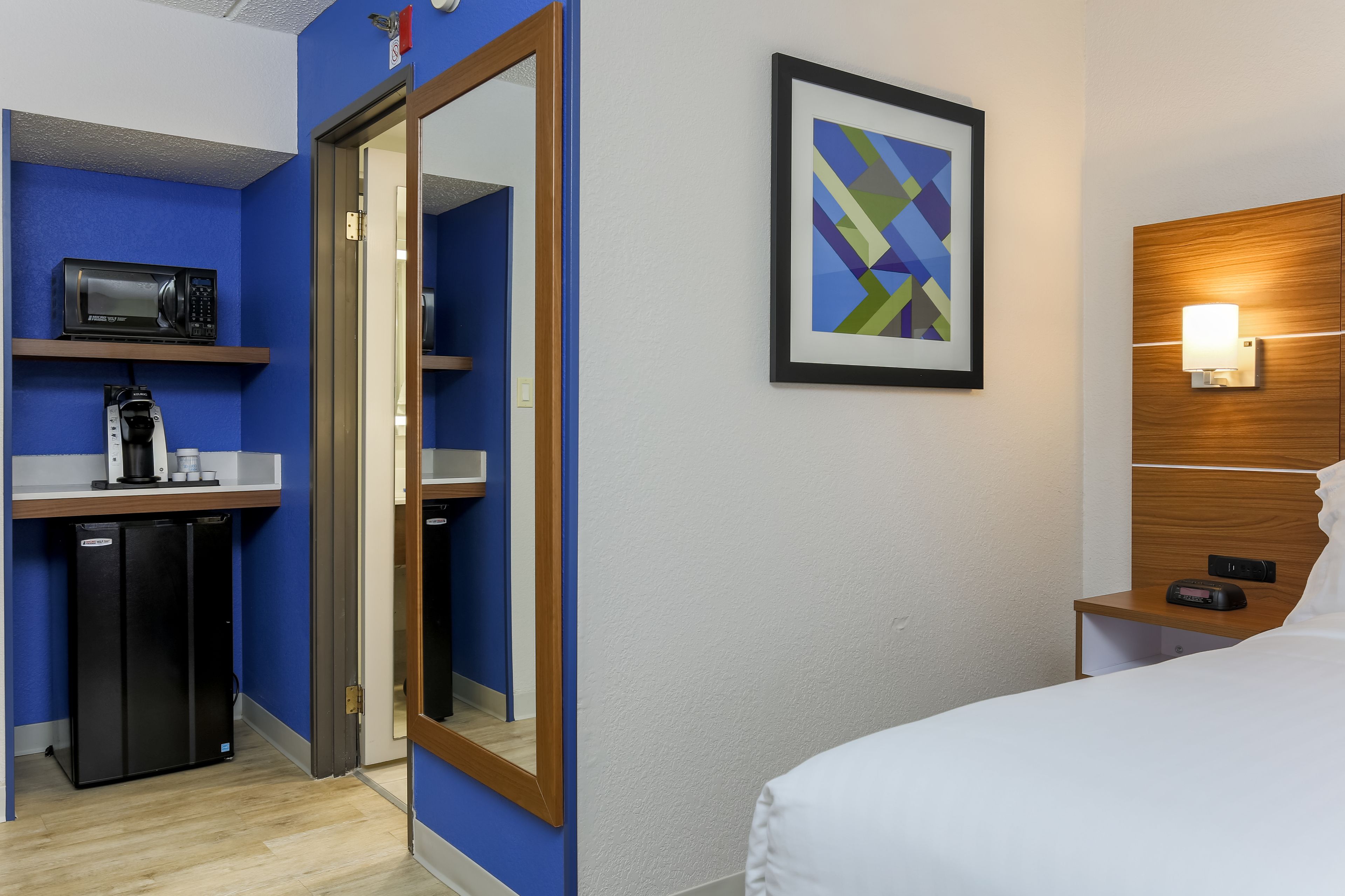 A hotel room with a bed, a bedside table with a lamp, a microwave, a refrigerator, and a framed abstract painting on the wall.