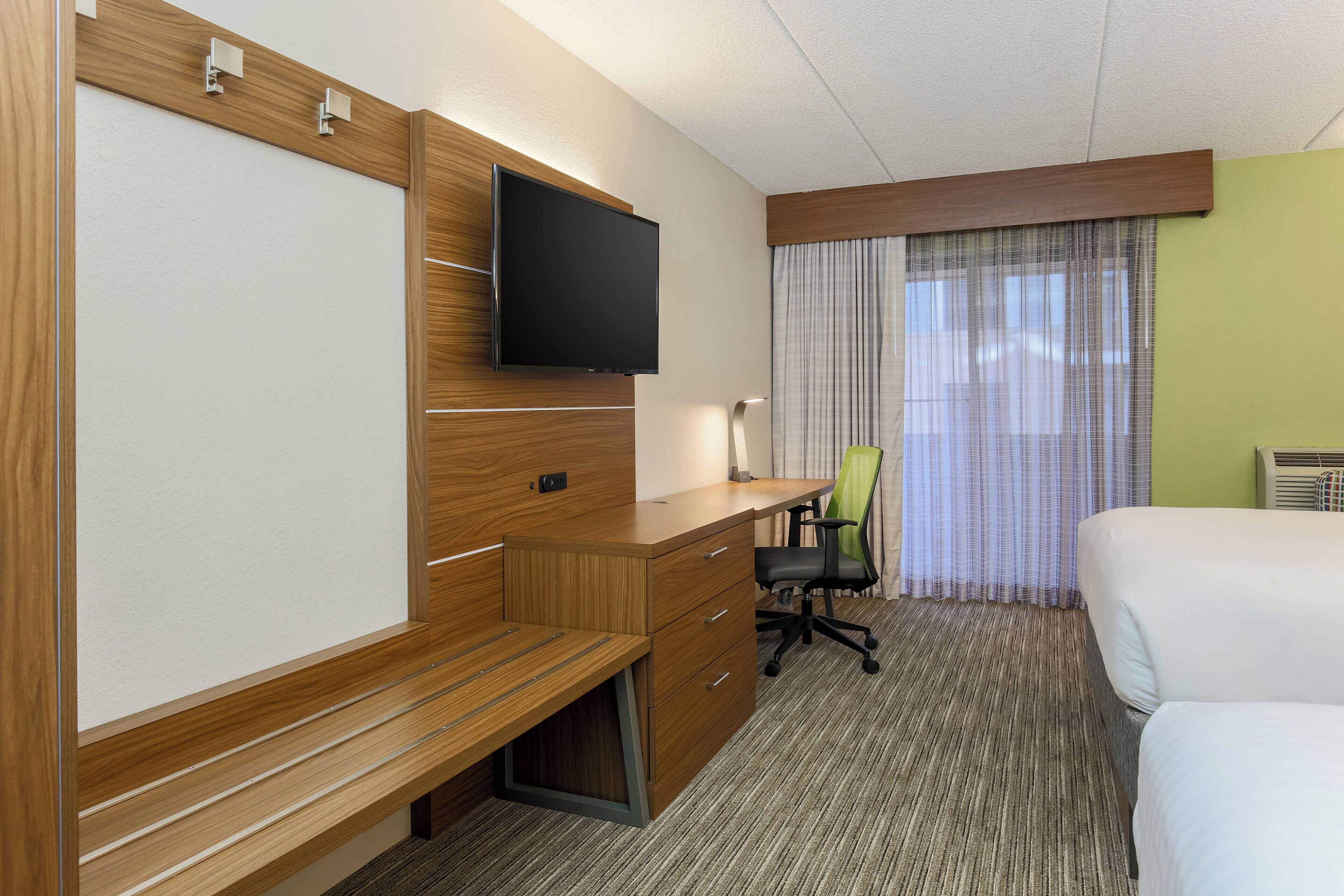A hotel room with a wooden desk, a flat-screen TV, a green office chair, a bed, and a window with curtains.