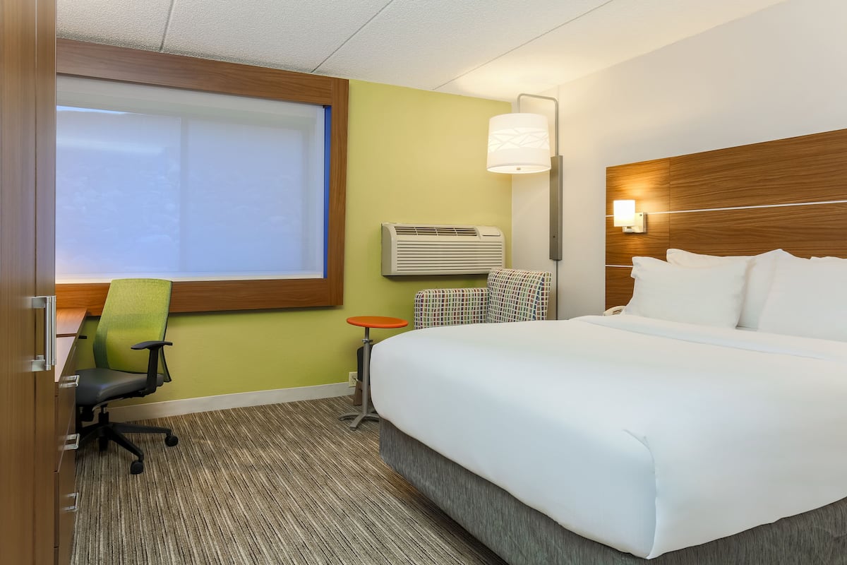 A hotel room with a large bed, a desk with a chair, an air conditioning unit, and a window with blinds.