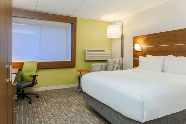 A hotel room with a large bed, a desk with a chair, an air conditioning unit, and a window with blinds.