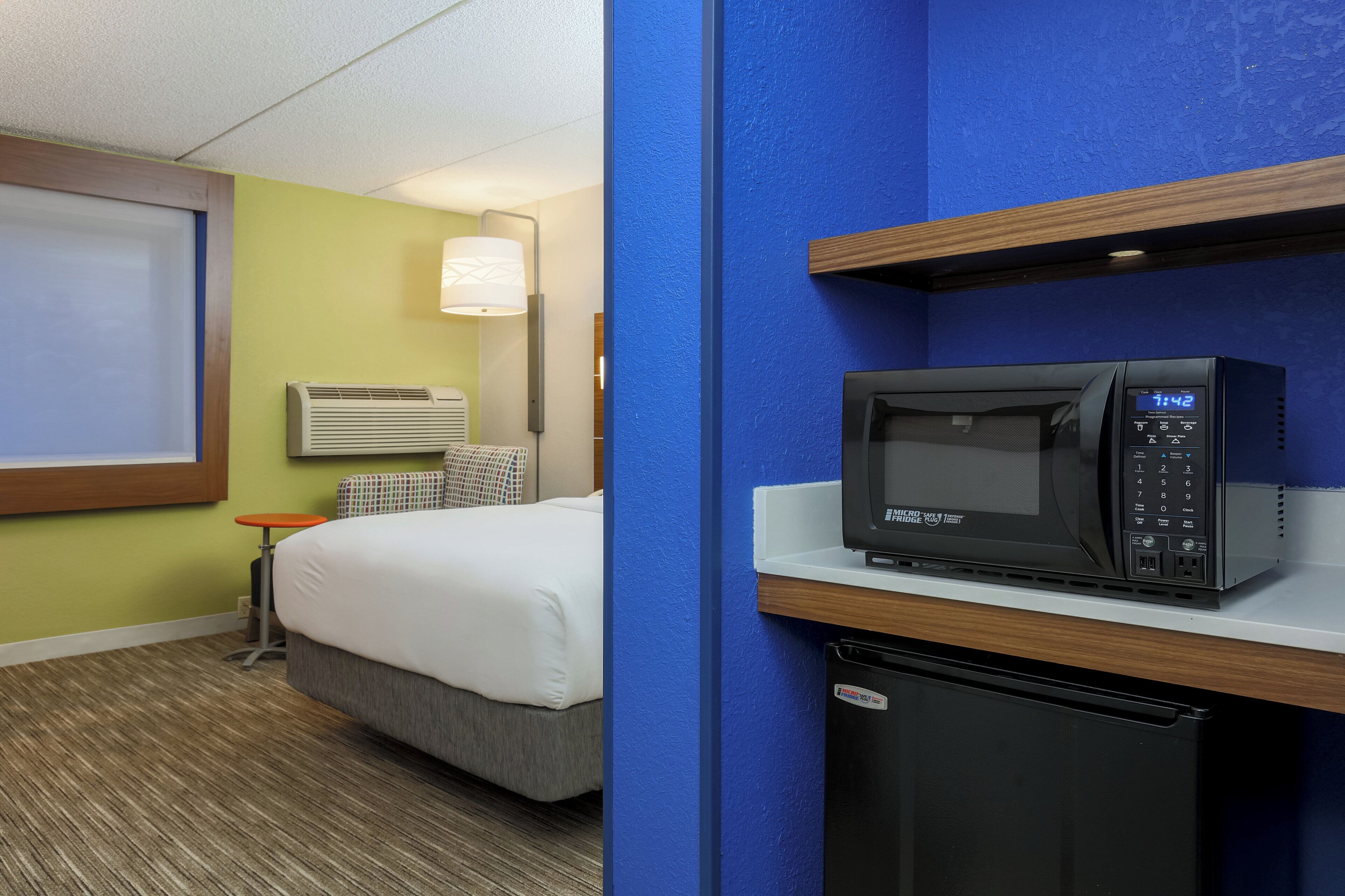 Quarto de hotel com cama, micro-ondas, mini-geladeira, mesa de cabeceira e luminária.