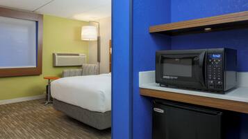 Quarto de hotel com cama, micro-ondas, mini-geladeira, mesa de cabeceira e luminária.