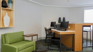 Une chambre d’hôtel avec un canapé vert, un bureau doté de deux écrans, une imprimante et une fenêtre.