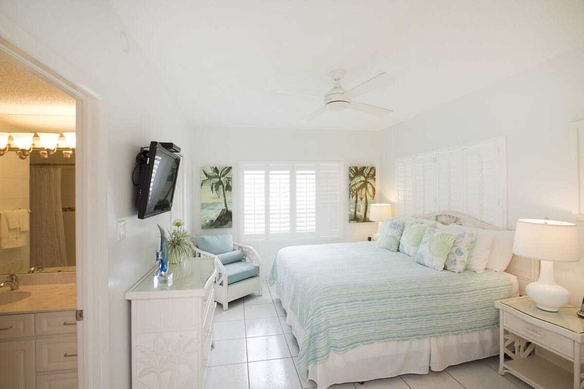 Quarto com cama, mesa de cabeceira, penteadeira e televisão fixada na parede.