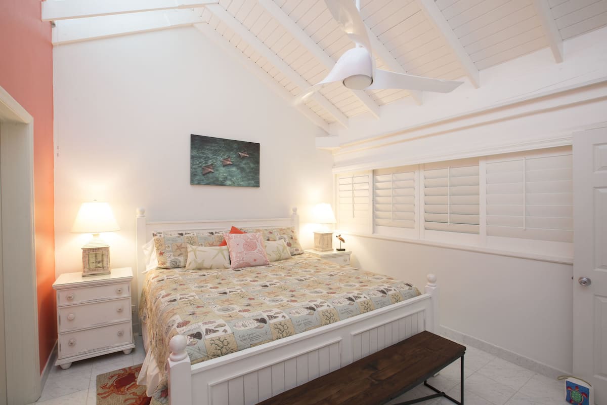 Quarto com cama, mesas de cabeceira, um banquinho, ventilador de teto e janela com persianas.