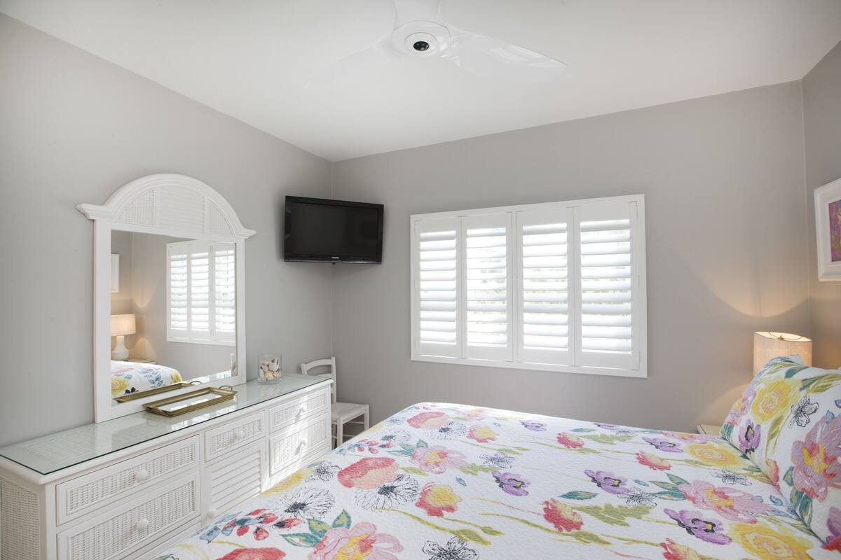 Quarto com cama, penteadeira, TV e janela com persianas.