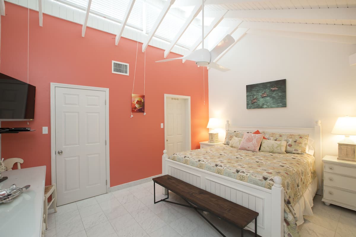 Quarto com cama, mesas de cabeceira, um banco, TV e ventoinha no teto.