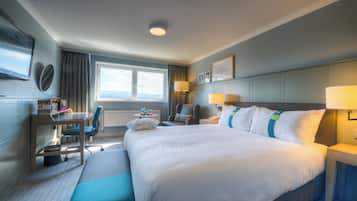 A hotel room with a large bed, a desk with a chair, a window with a view, and a flat-screen TV mounted on the wall.
