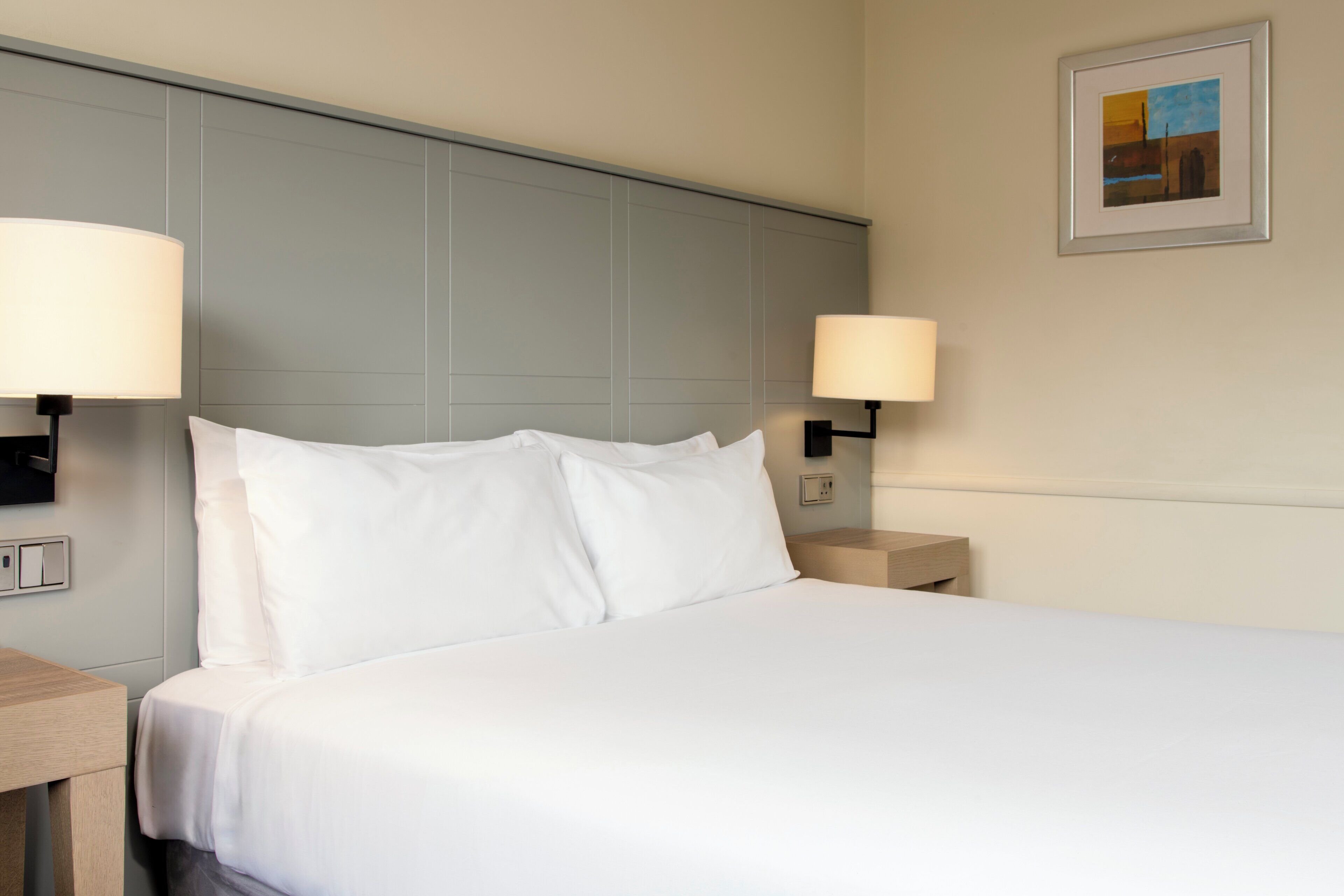 A neatly made bed with white linens in a hotel room, two bedside lamps, and a framed picture on the wall.
