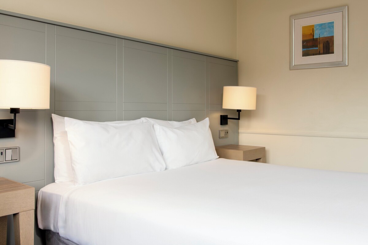 A neatly made bed with white linens in a hotel room, two bedside lamps, and a framed picture on the wall.