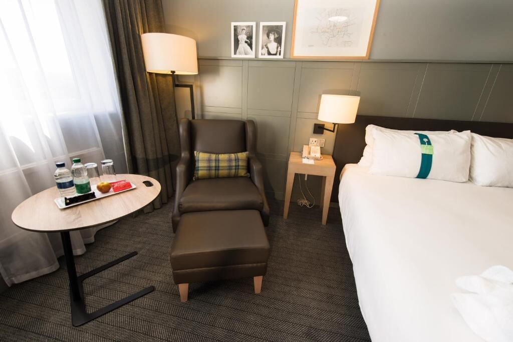 A hotel room with a bed, a chair, a small table with a drink and snacks, and a lamp.