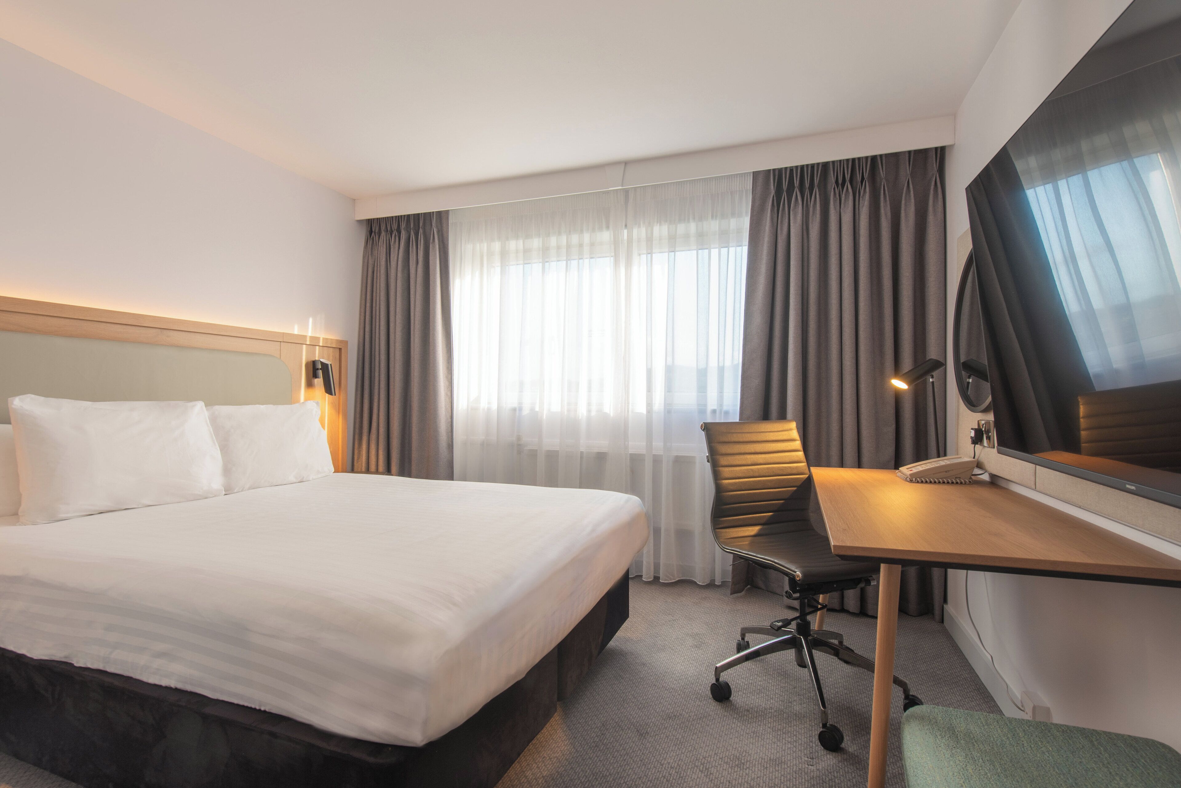 A hotel room with a large bed, a desk, a chair, a TV, and a window with curtains.