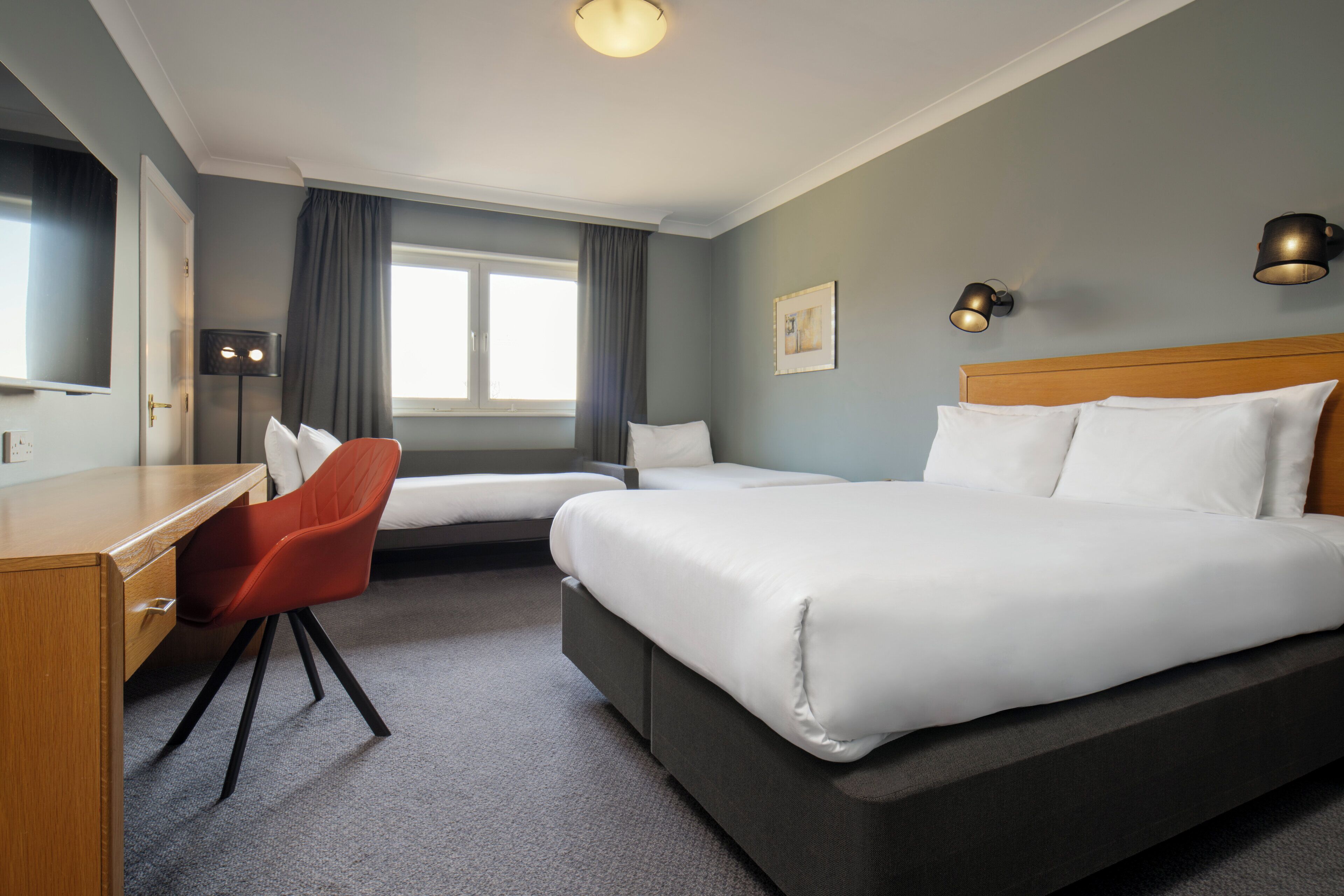 A hotel room with a large bed, a desk with a chair, a window with curtains, and two wall-mounted lamps.