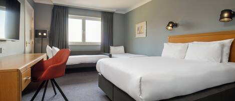A hotel room with a large bed, a desk with a chair, a window with curtains, and two wall-mounted lamps.