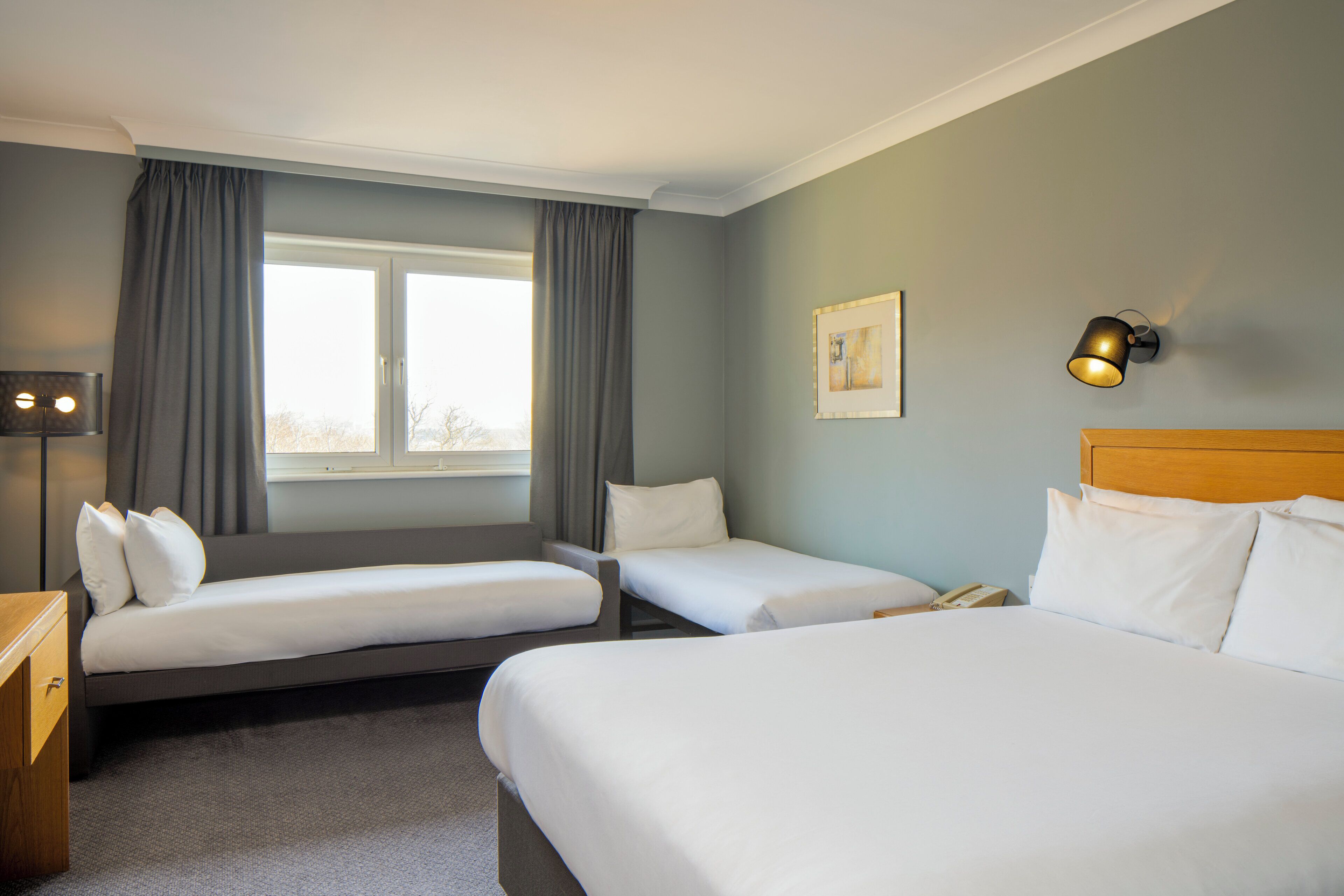 A hotel room with two single beds, a larger bed, a bedside table with a lamp, a window with curtains, and a wall-mounted light fixture.