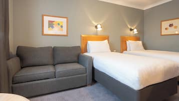 Una habitación de hotel con un sofá gris, dos camas con ropa blanca y dos lámparas fijadas a la pared.