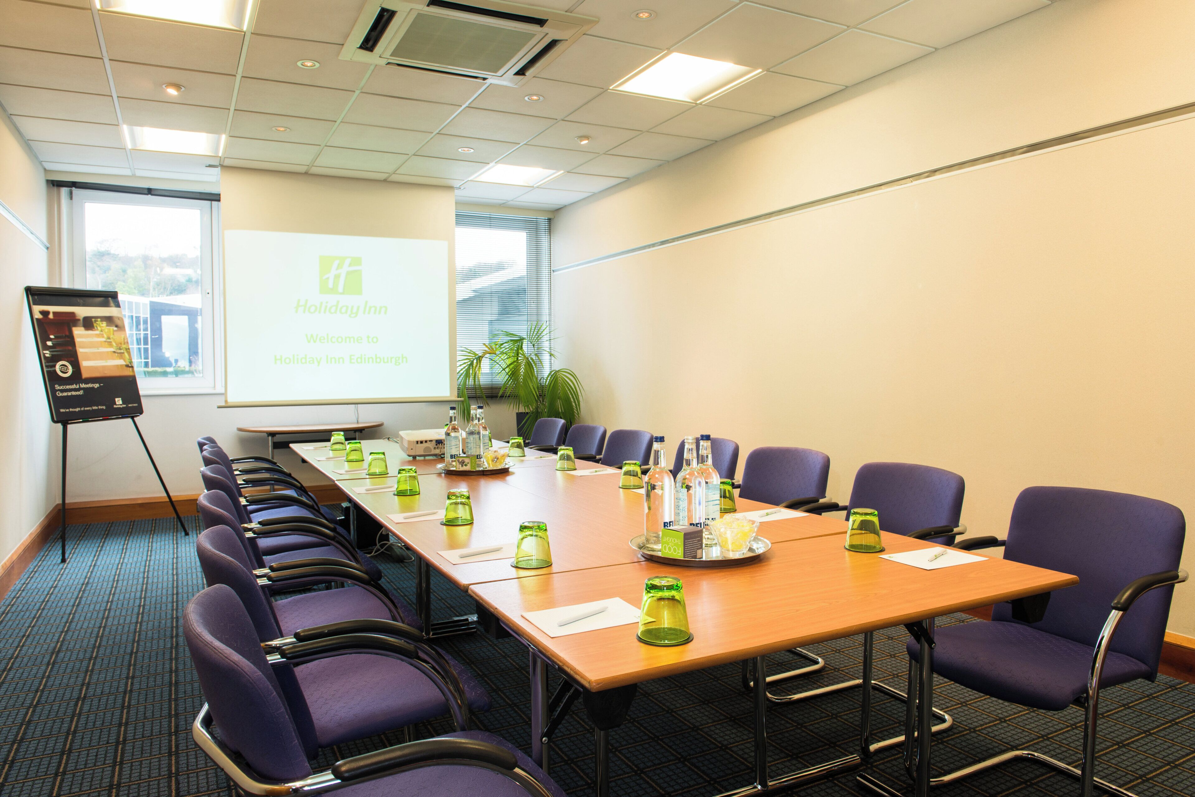 A conference room with a long table, chairs, a projector screen, and a presentation about Holiday Inn Edinburgh.