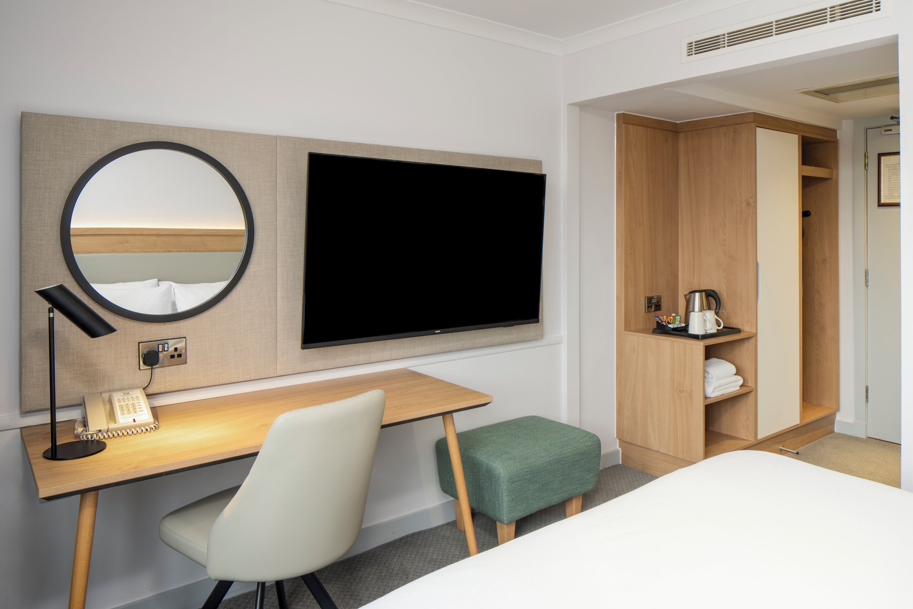 Une chambre d’hôtel comprenant un lit, un bureau avec une chaise, une télévision, un miroir, une armoire et un petit tabouret vert.