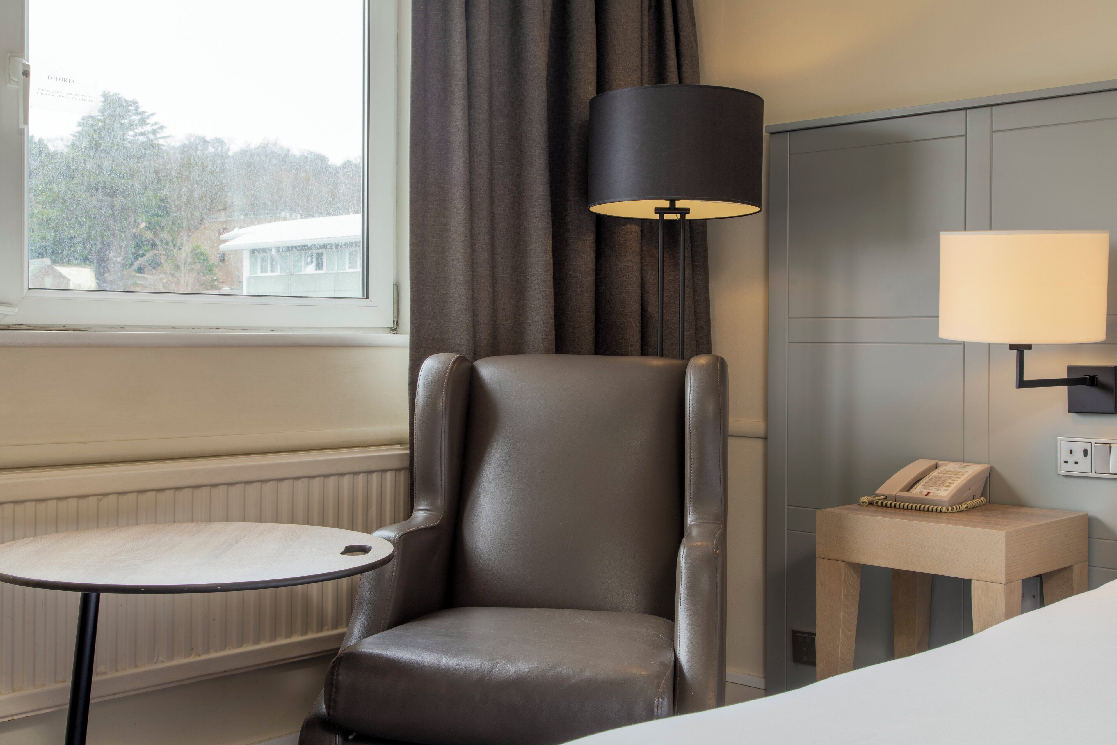 A hotel room with a leather armchair, a small round table, a lamp, and a bedside table with a phone.