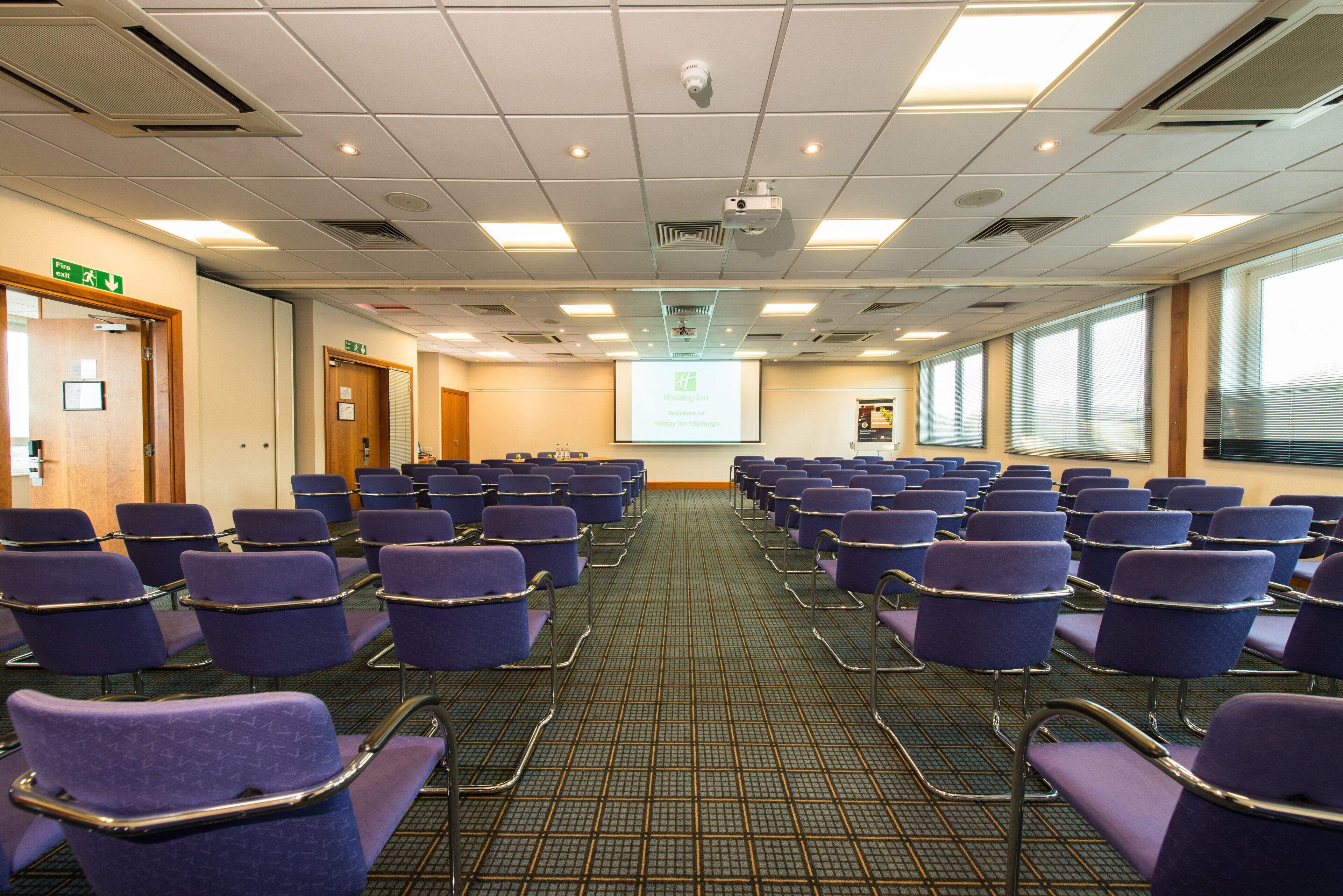 A conference room with rows of purple chairs, a projector screen, and a fire exit sign.