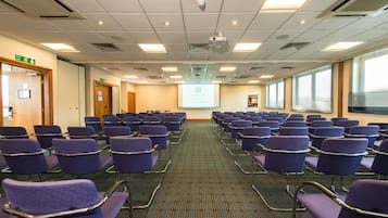 A conference room with rows of purple chairs, a projector screen, and a fire exit sign.