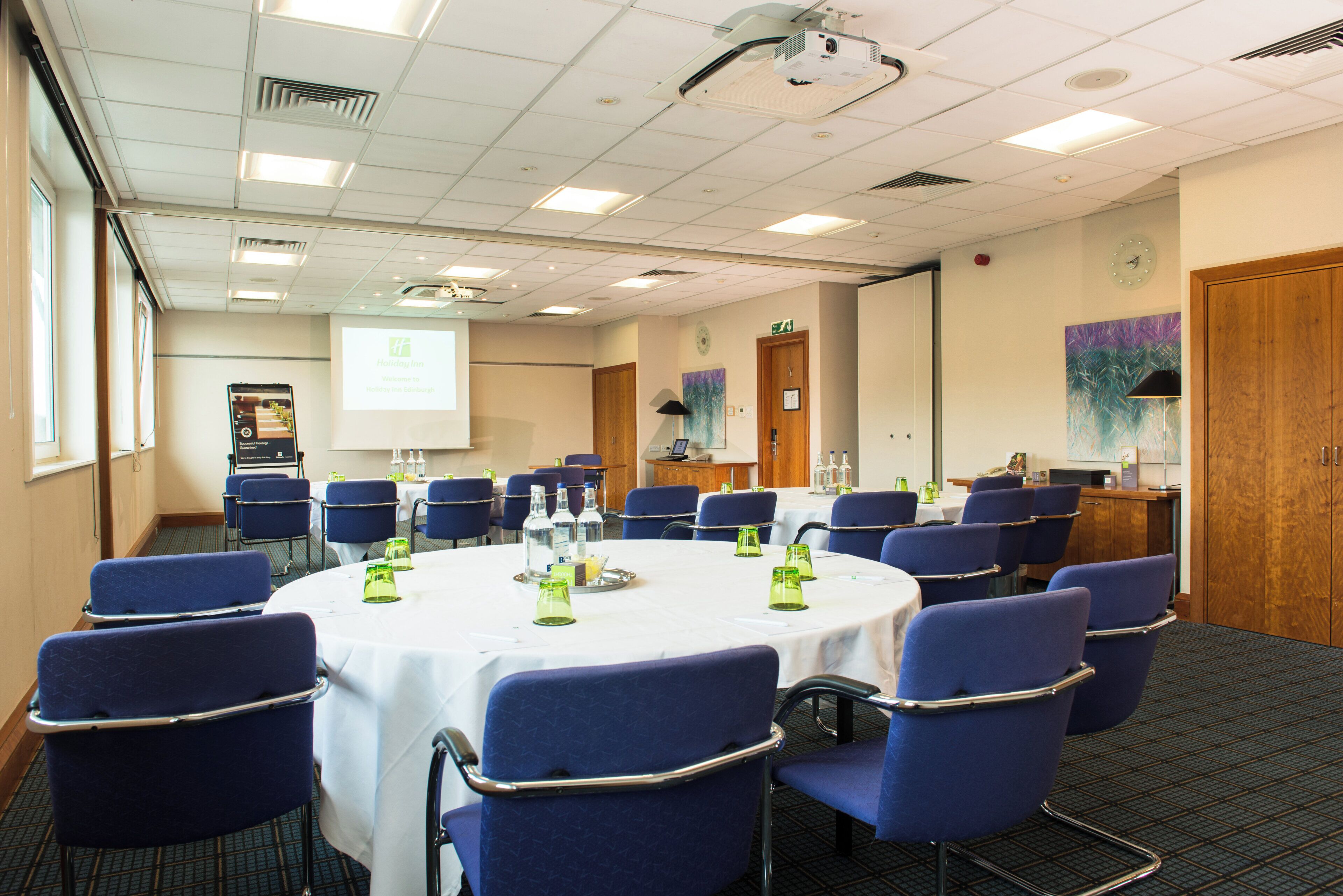 A conference room with a round table, blue chairs, and a projector screen.