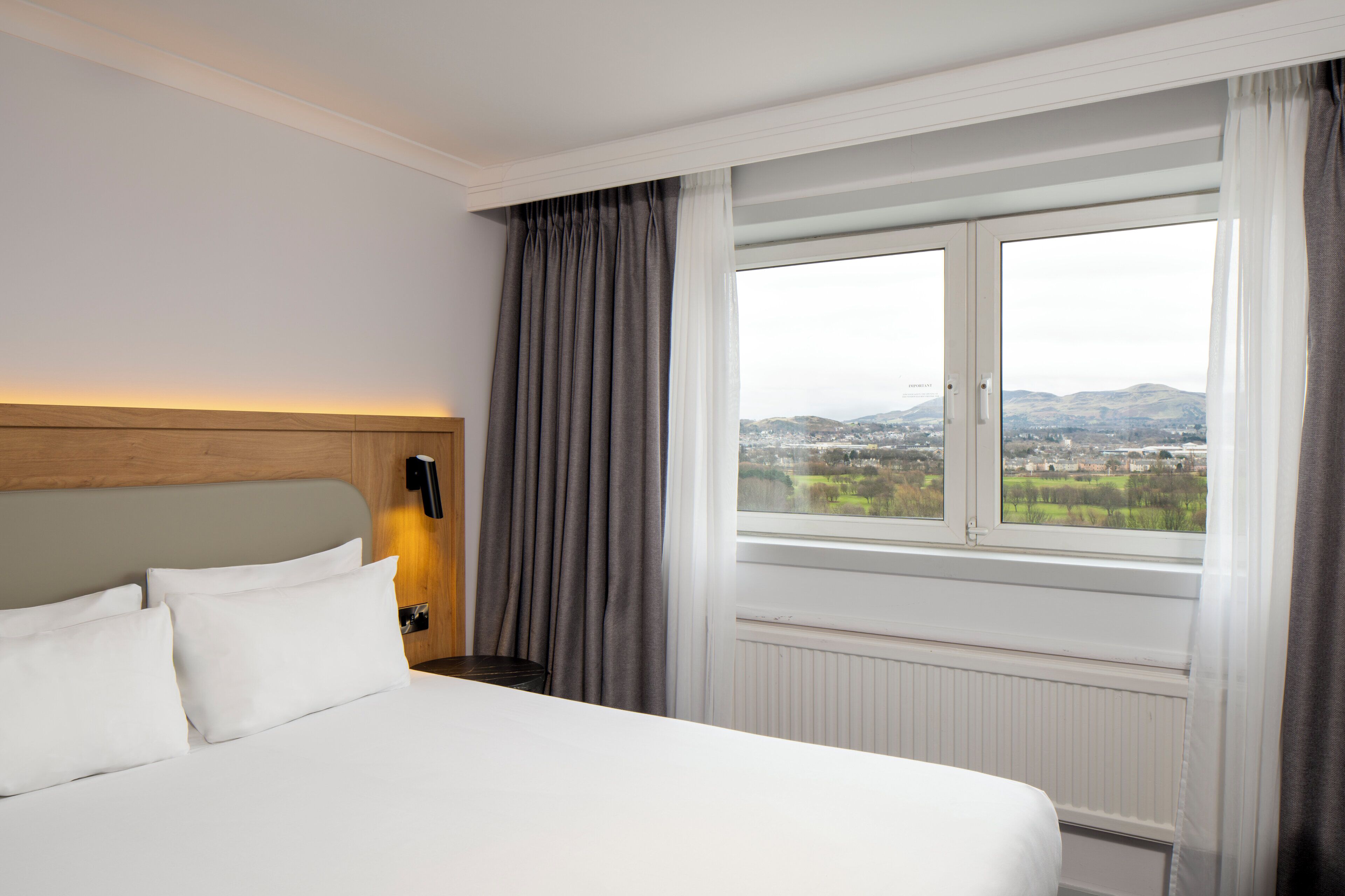 A hotel room with a large bed, a wooden headboard, a nightstand with a phone, and a window with curtains.