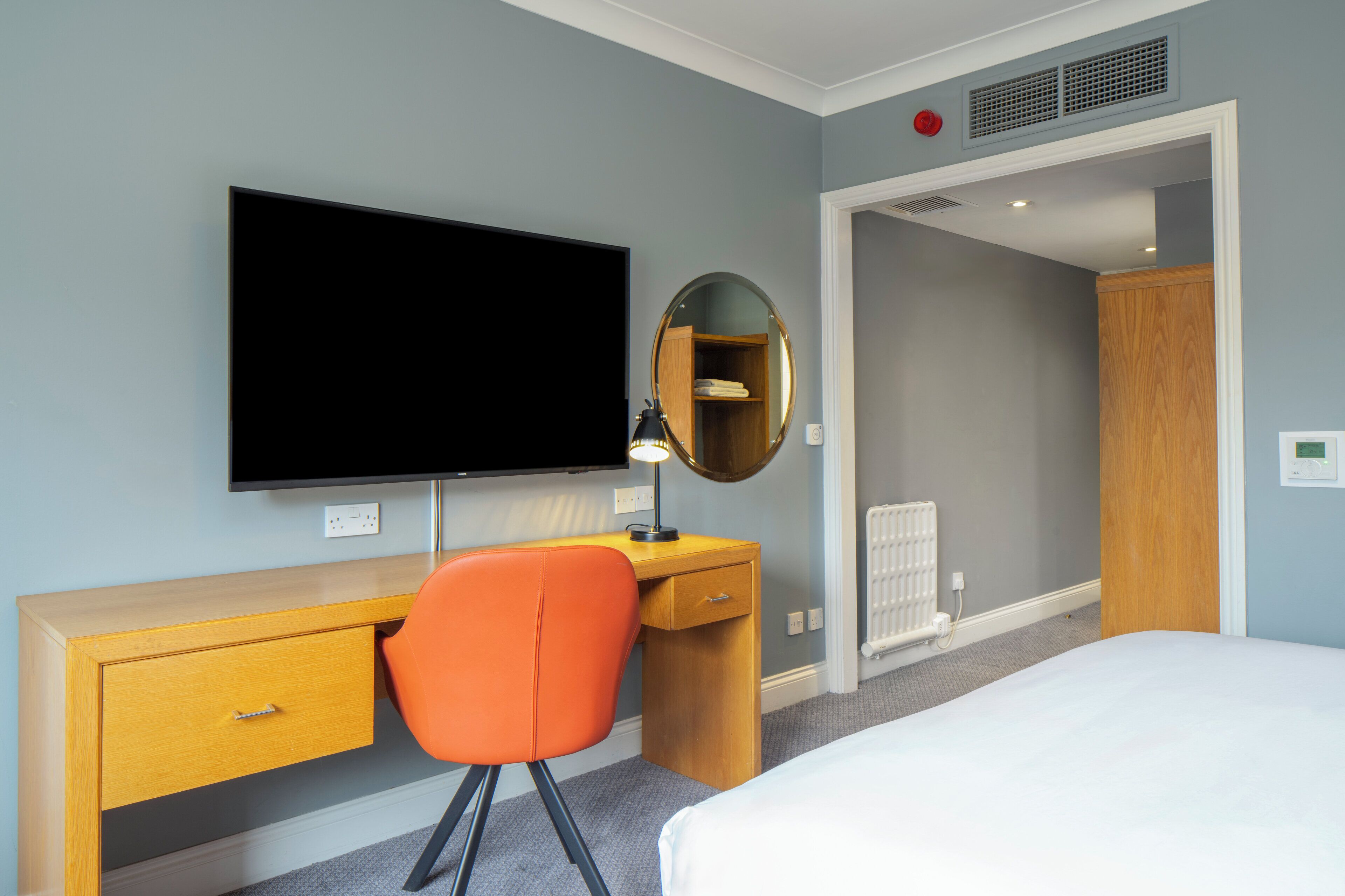 A hotel room with a bed, a desk with a chair, a television, a mirror, and a wardrobe.