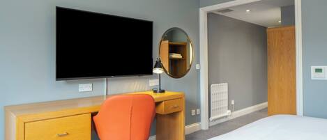A hotel room with a bed, a desk with a chair, a television, a mirror, and a wardrobe.