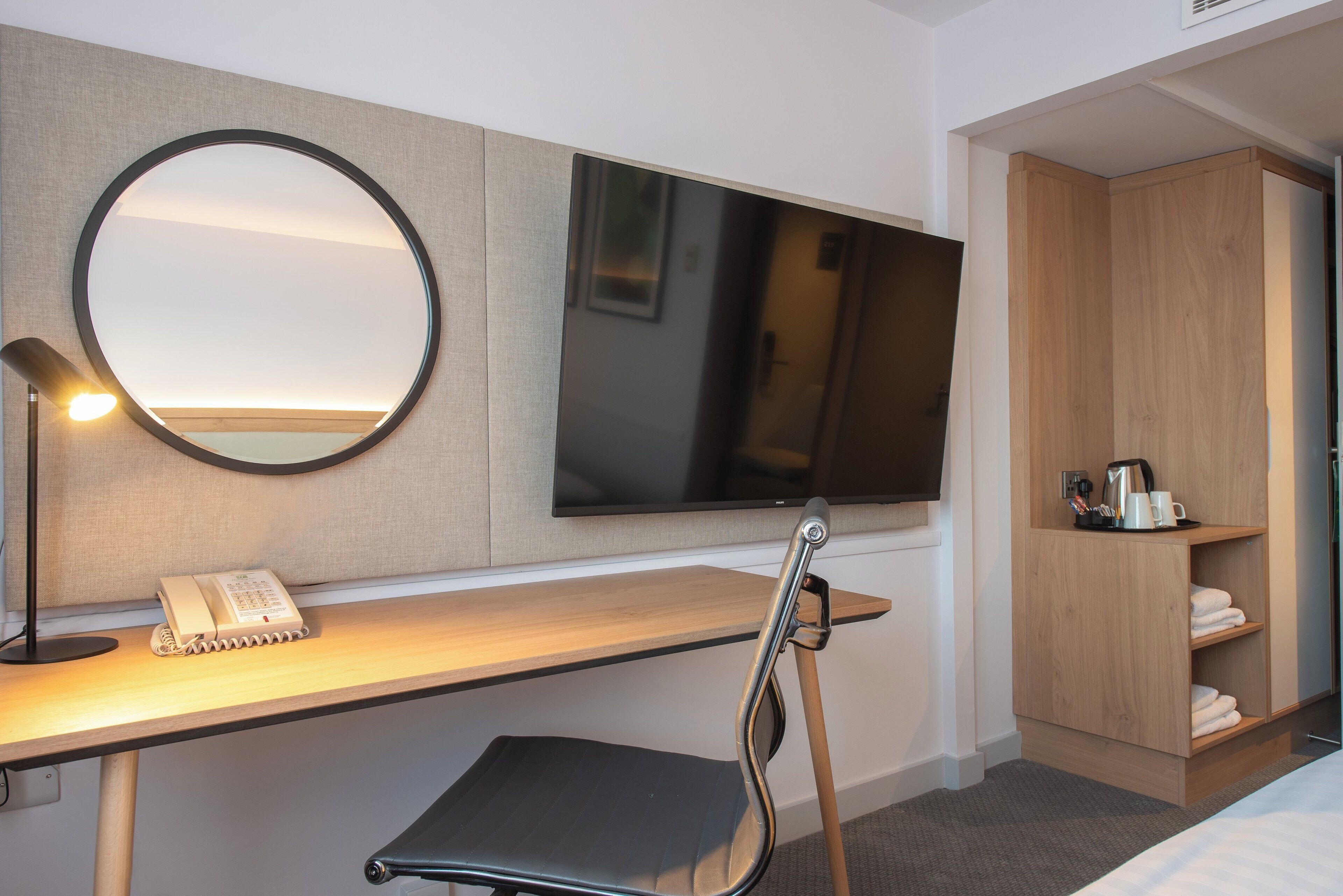 Habitación de hotel con escritorio, silla, espejo redondo, televisor de pantalla plana y armario.