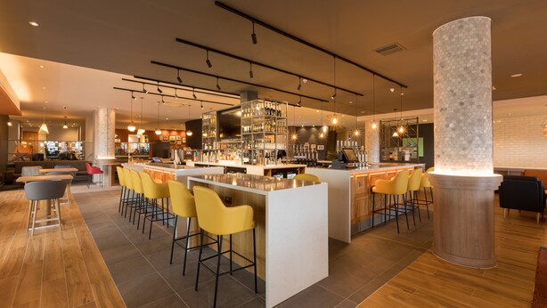 Un espace bar moderne doté d’un îlot central, de tabourets de bar et d’une variété de bouteilles et de verres disposés sur des étagères.