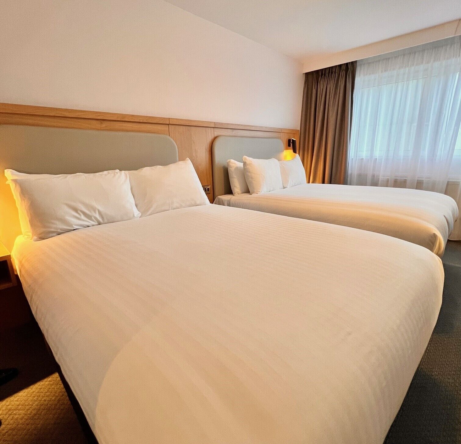 A hotel room with two beds, a wooden headboard, a bedside table, and a window with curtains.