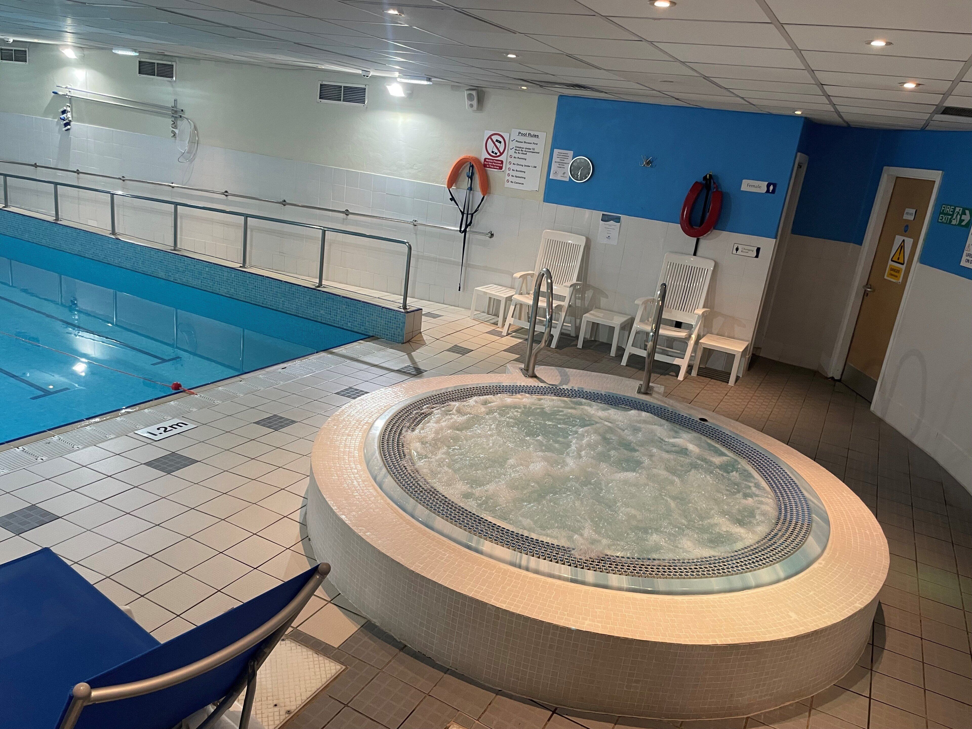 A swimming pool with a hot tub, tiled flooring, and white chairs.