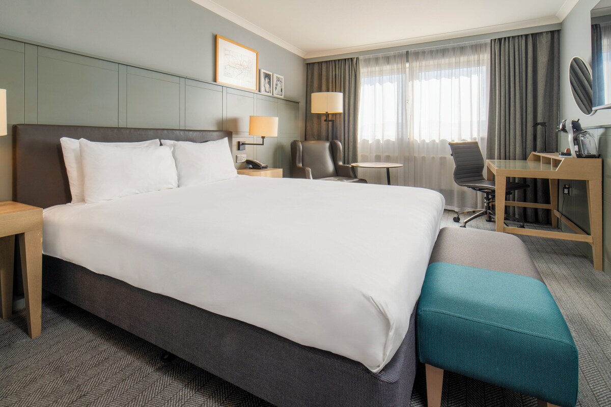 A hotel room with a large bed, a desk, a chair, a small table, and a bench.