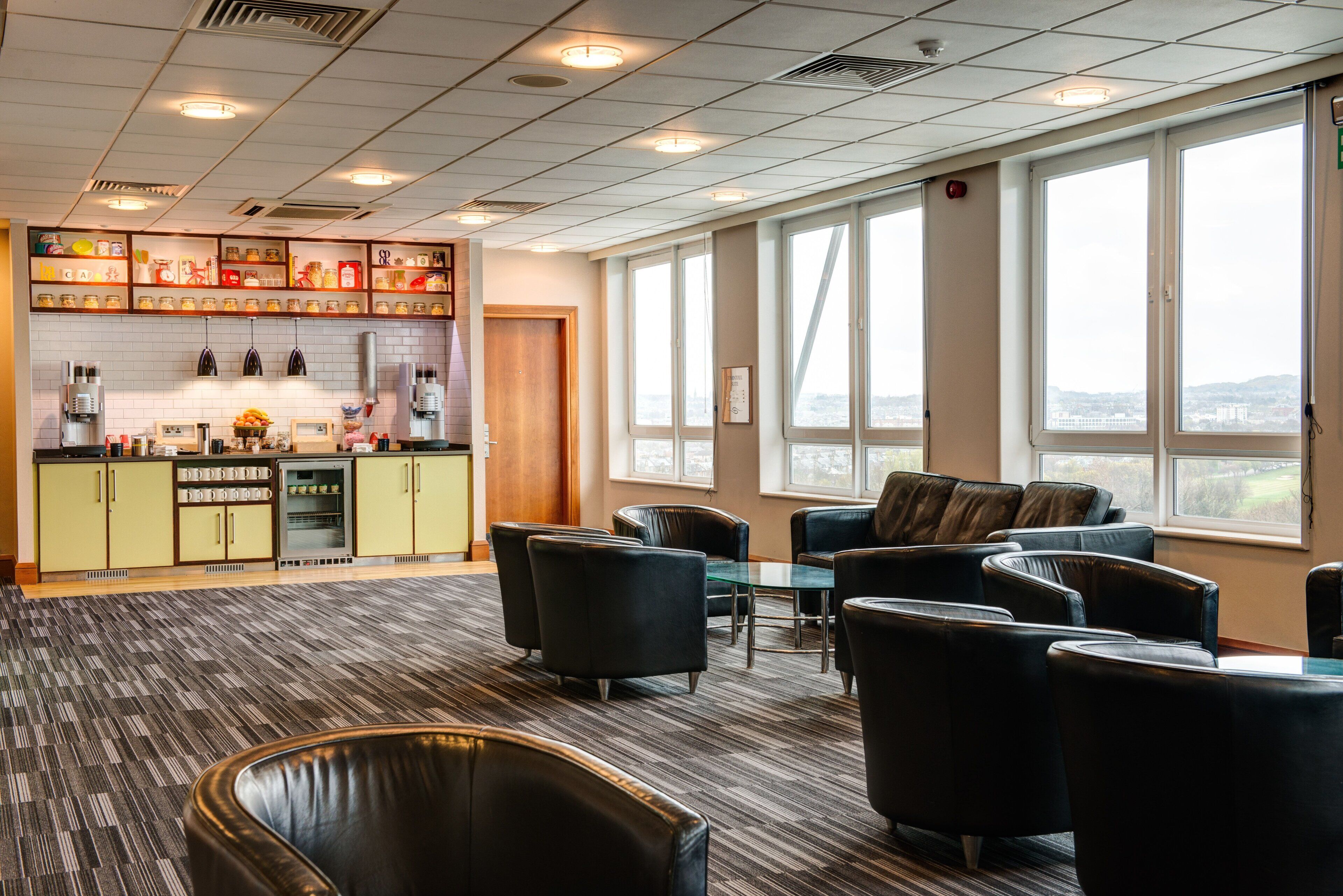 A modern kitchen with a coffee machine, refrigerator, and shelves with jars. There is a seating area with leather chairs and a view of the city through large windows.