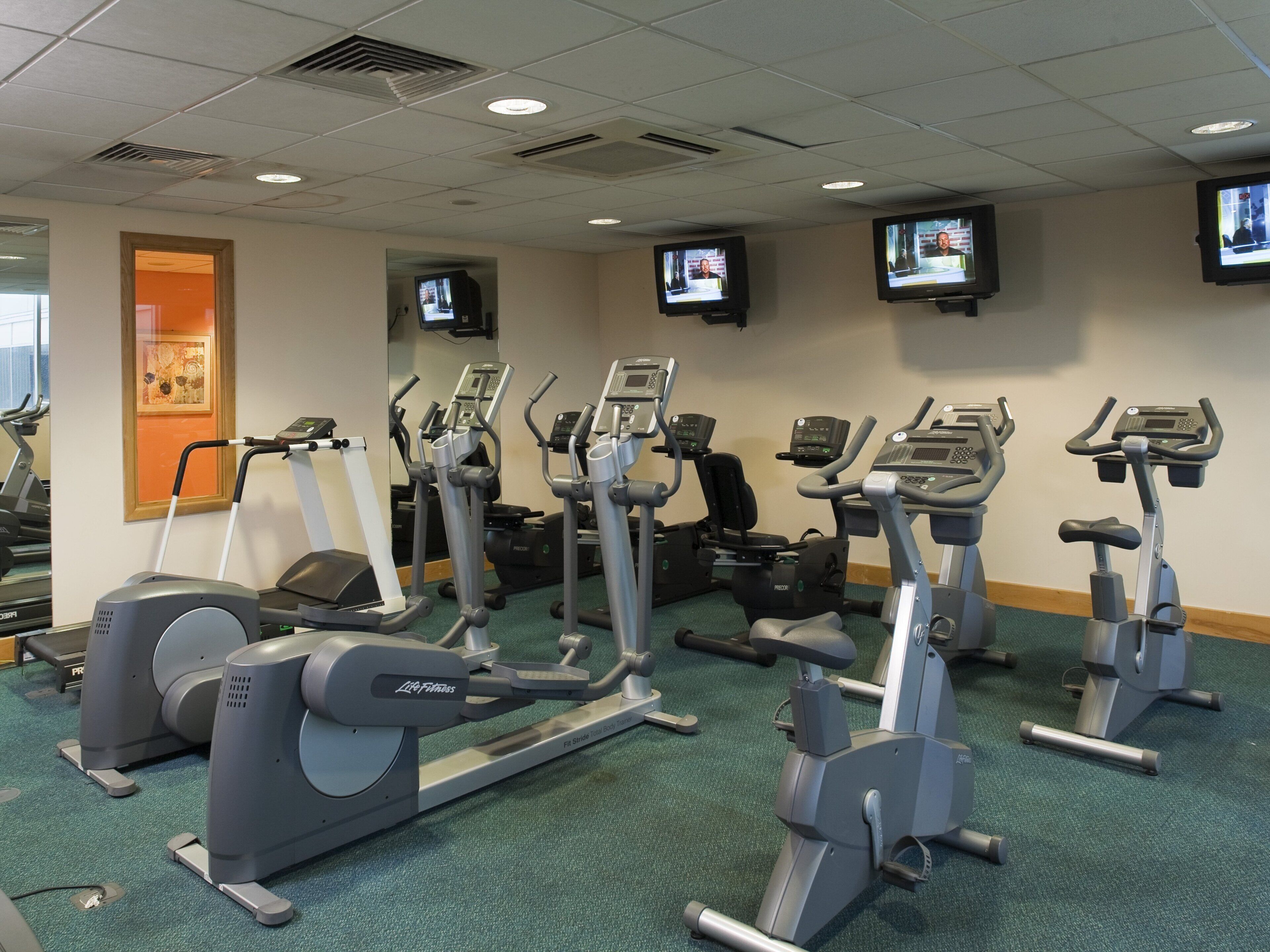 A gym with multiple cardio machines, including treadmills and ellipticals, and mirrors on the walls.