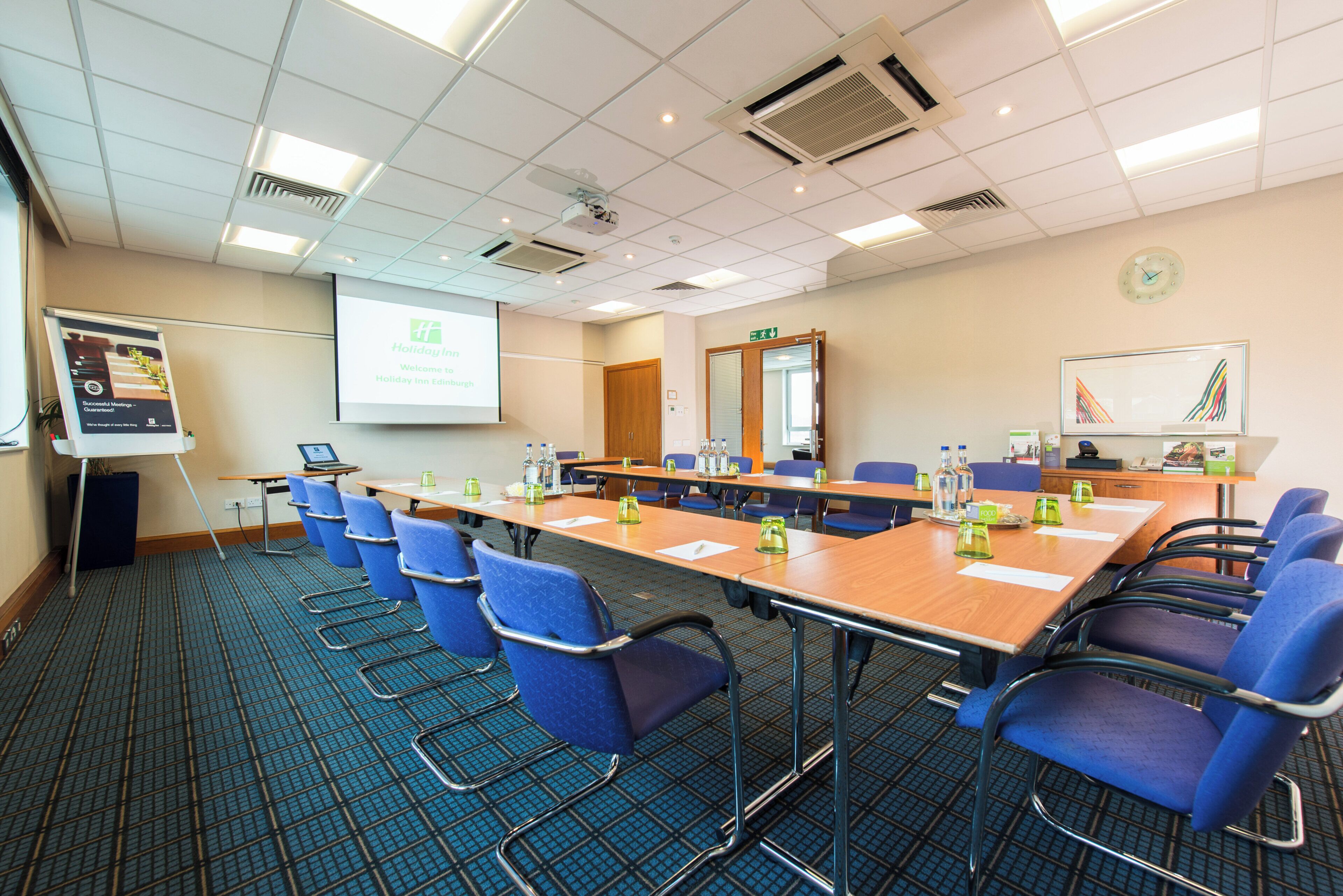 Ein Konferenzraum mit einem langen Tisch, blauen Stühlen, einer Projektorleinwand und einer Uhr an der Wand.