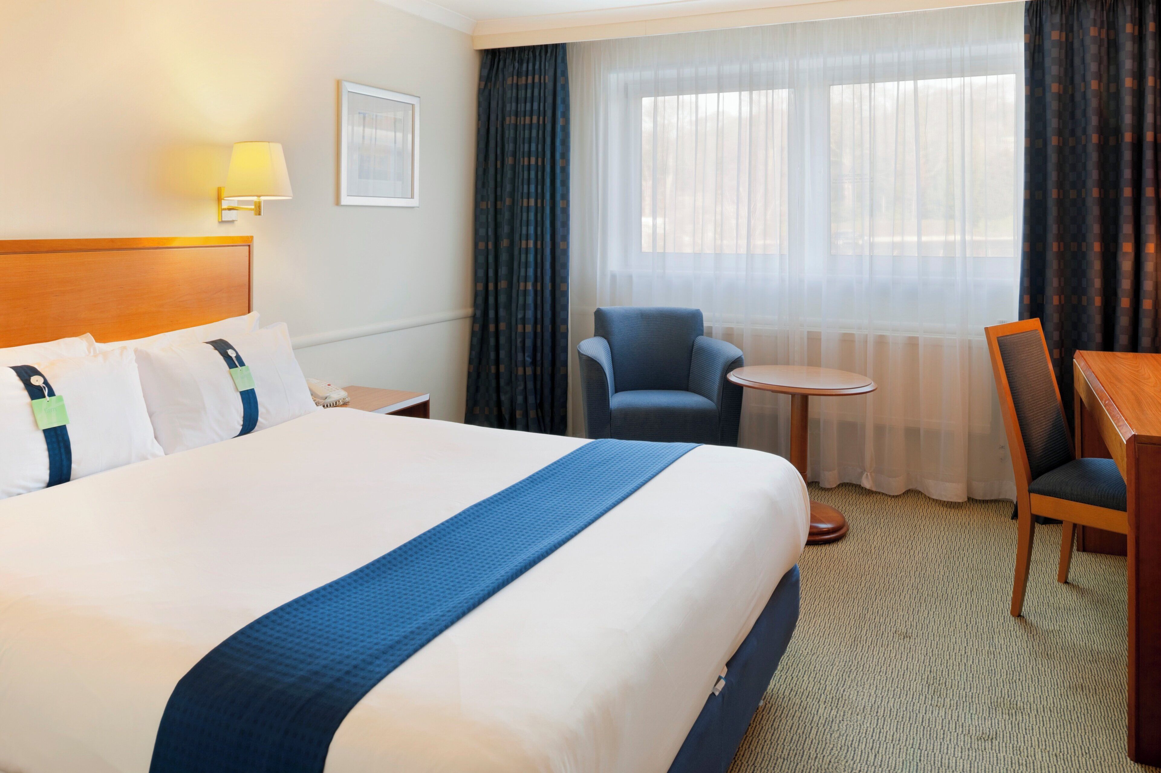 A hotel room with a large bed, a small round table, two chairs, a desk, a lamp, and a window with curtains.