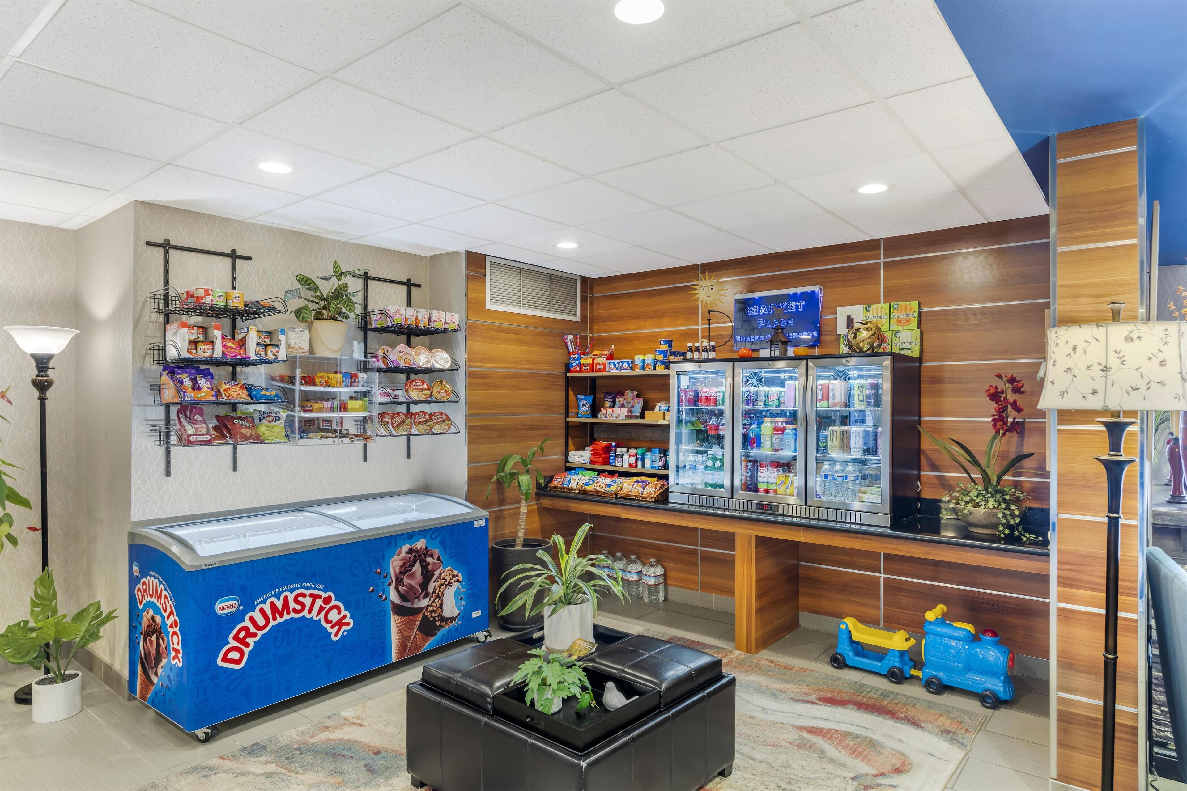 Een hotel lobby met een Drumstick ijskoeler, een koelkast met snacks, een speeltreintje en een potplant.