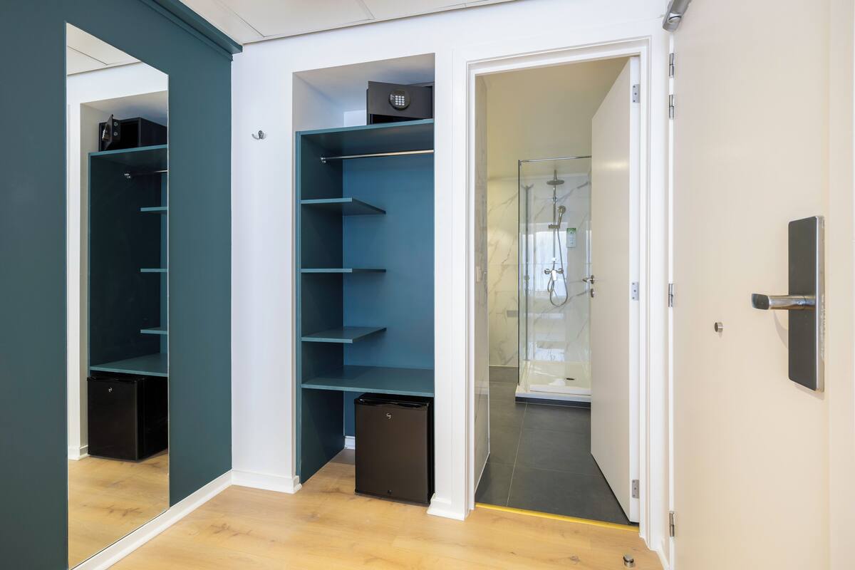 A modern bathroom with a glass shower enclosure, a white door, and a teal-colored shelving unit with a black trash bin.